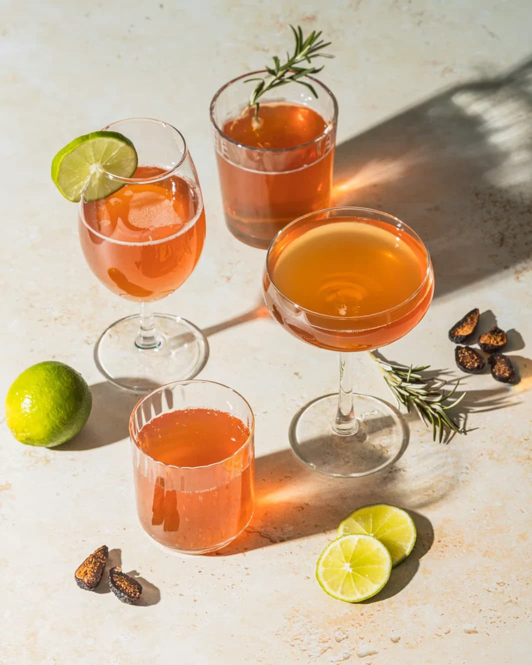 Four glasses of amber drinks with lime and rosemary on a sunlit table
